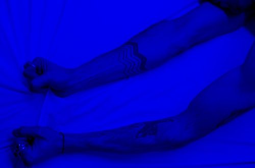 Close-up of tattooed arms grasping a white bedsheet under blue lighting, depicting anxiety and tension.